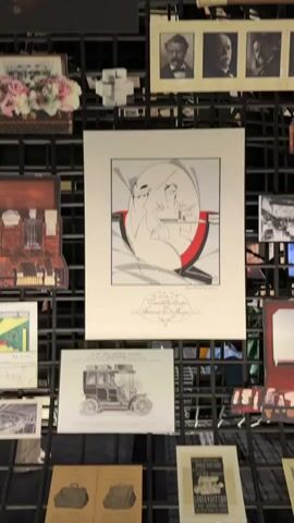 The video shows a wall display of vintage Louis Vuitton items. The display is organized on a black grid background. In the center of the frame is a large framed illustration of two figures in a stylized art deco design. Below this illustration is a framed image of a vintage automobile. To the left of the central illustration, there are smaller images of Louis Vuitton products and a framed arrangement of pink and white flowers. Above the central illustration, there are four framed portraits of men and several canes with decorative handles. In the upper left corner, there is an image of an ornate Louis Vuitton trunk. Other items visible include vintage advertisements, logos, and product displays. The lighting suggests an indoor exhibition or museum setting.