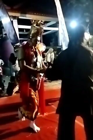 The video shows an outdoor nighttime performance. A person in elaborate traditional costume and headdress is dancing on a red stage. The costume is red and gold. In the background, other people are watching the performance, some sitting and some standing. There are lights illuminating the stage and some decorative balloons are visible. The overall environment appears to be festive.