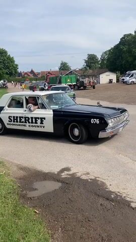 A black and white vintage sheriff's car is driving slowly on a dirt road. The car has "SHERIFF" and "MOONSHINE COUNTY" written on the side. The number "076" is on the front fender. Inside the car, a man wearing sunglasses and a striped shirt is driving. Two women are in the passenger seat. In the background, there are trees, buildings, a bus, and other vehicles. The sky is cloudy.