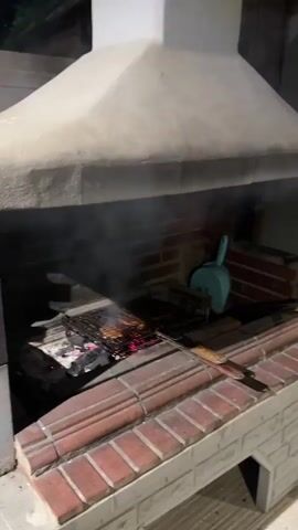 The video shows a person grilling meat over hot coals in an outdoor kitchen setting. The grill is made of brick and has a large concrete hood. In the background, a kitchen area is visible with cabinets, a refrigerator, and a coffee machine. Two people are in the kitchen area, one of whom appears to be preparing food. The overall scene suggests a casual gathering or meal preparation.