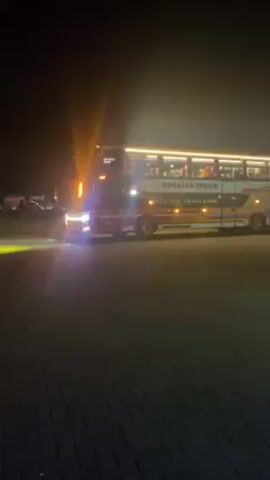 At night, the camera pans across a brightly lit parking lot containing several double-decker buses. The first bus is red, white, and yellow and has the words “Rosalia Indah” on the side. The second bus is mostly white with blue and green decorative lights and two people standing near the front.