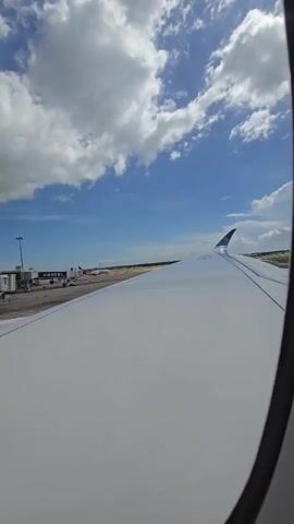 The video shows a view from an airplane window as the plane is on the tarmac at an airport. The wing of the plane is visible in the foreground, taking up most of the frame. Beyond the wing, the airport runway is visible, along with several jet bridges connected to terminals. Other airplanes are parked at the terminals. The sky is blue with fluffy white clouds. The sun is shining brightly. The camera appears to be stationary, providing a static view of the scene.