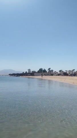 The video captures a serene beach scene in Safaga, Egypt, on a bright, clear day. The calm, shallow, turquoise water of the sea, revealing the sandy seabed with gentle ripples, dominates the foreground. In the background, a sandy beach stretches along the shore, lined with resort-style buildings, thatched umbrellas, and a few palm trees.

At the beginning of the video, a single person is seen walking leisurely along the distant shoreline. As the camera pans slightly to the left, two individuals are visible relaxing on blue floating mats in the tranquil water. Majestic mountains frame the distant horizon across the water. The sky above is a brilliant, cloudless blue, indicating sunny weather. The overall ambiance is one of peace and relaxation, typical of a vacation destination.