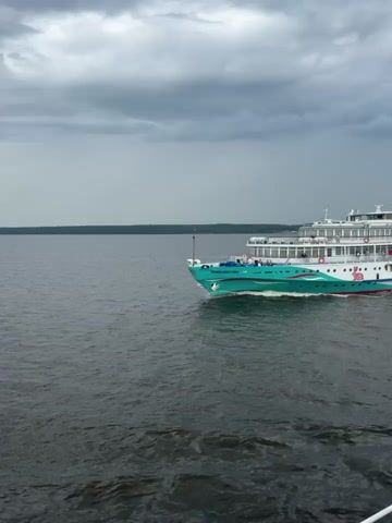 The video shows a white and teal cruise ship moving across dark water under a cloudy sky. The ship has multiple decks with windows and railings. People are visible on the decks, some standing near the railings, and some appear to be waving. The ship has a large teal and white design along its side, including a stylized pink rabbit image. The water in the foreground is choppy and dark. The horizon line is visible in the distance, with a faint line of trees or land. The clouds are heavy and dark, suggesting overcast weather.