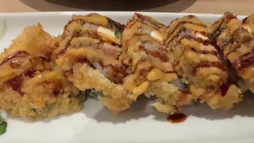 The video shows a plate of sushi rolls on a white plate. The sushi rolls are deep-fried and drizzled with sauce. They are arranged in a circle around a pile of shredded daikon radish. There is also a small mound of wasabi and a few slices of pickled ginger on the side. The plate is on a wooden table.