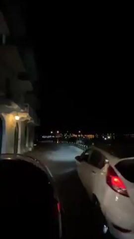 At night, a car exits a parking space next to a white car. The road has a row of parked cars on each side. The road leads to a view of a lit-up building on a hill, across an expanse of other buildings. There is a brief honk of the car's horn. The building on the hill is likely a large historical structure. The environment is a city near a body of water.