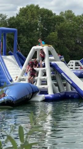 The video shows an inflatable water park on a lake. Several children and young adults are on the inflatable structures. One child is on a blue slide, and another is in the water. Multiple people are on the stairs of a white and blue inflatable structure. One person is at the top of the structure, and others are on the steps. A blue inflatable object is in the water, and a person is lying on it. Trees and a cloudy sky are in the background.