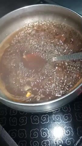 A stainless steel pot filled with a dark brown liquid, likely a broth or sauce, is shown bubbling vigorously. The liquid is filled with numerous small bubbles, indicating it is at a rolling boil. Some larger, reddish-brown pieces of food are visible suspended in the liquid, along with smaller, lighter-colored pieces, possibly spices or herbs. A utensil, appearing to be a metal slotted spoon or strainer, is partially submerged in the liquid on the right side of the pot, possibly used for stirring or removing ingredients. The pot is sitting on a dark, patterned stovetop. The lighting is overhead and casts a bright reflection on the stovetop. The focus is on the boiling liquid and the contents within the pot.