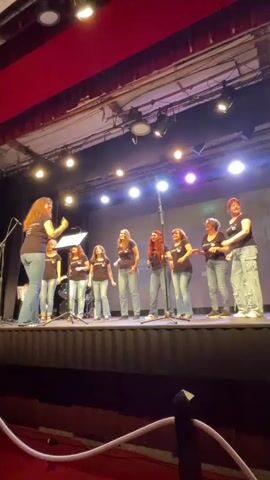A group of nine females, all wearing dark t-shirts and denim jeans, stand on a stage. One woman, presumably the director, stands to the left, facing the group and gesturing with her left hand. The other eight women are arranged in a line across the stage, facing forward, and appear to be singing. They are holding microphones. The stage is lit by overhead spotlights and appears to be in a theater or auditorium setting, indicated by the red curtain at the top of the frame and the seating area in the foreground.