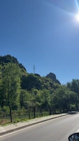 The video is captured from a moving vehicle on a sunny day. The road runs along the right side of the frame, with a sidewalk in front of it. To the left of the road, there is a lush green hillside covered with trees and vegetation. In the distance, a large rock formation or mountain peak is visible, topped with a man-made structure, likely a power line tower. The sky is a clear, bright blue, with the sun shining intensely in the upper right corner, creating lens flare. A wooden fence and some scattered wooden posts are visible along the edge of the hillside, bordering the sidewalk. The overall impression is a scenic drive through a natural landscape.