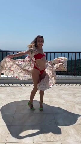 A woman in a red bikini and neon green heels stands on a tiled balcony overlooking a body of water and lush green hills. She is wearing a flowing, sheer kimono with a floral print. She turns around, the kimono billowing around her, and then spins back towards the camera, smiling. The video ends with her twirling, the kimono creating a large circle around her.