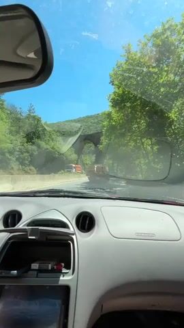 The video captures a first-person perspective from inside a car driving on a road. The driver is approaching a large, arched stone bridge or viaduct that spans over the road. The bridge is made of rough-hewn stones and is partially covered by lush green trees and vegetation on either side.

The road appears to be a highway or a well-maintained road, with a concrete barrier separating it from the embankment on the left. There are other vehicles on the road, including a red and white truck visible ahead and another orange truck further in the distance. The surrounding environment is dominated by dense, green trees, suggesting a rural or mountainous setting. The sky is a clear, bright blue, indicating a sunny day.

The dashboard of the car is visible, showing a car stereo with a digital display that reads "DX ST" and "99.90" in large font, along with other radio frequency information. There is also a slot with what appears to be a packet of cigarettes and a lighter inside. A rear-view mirror is also visible in the upper left corner of the frame.

The overall impression is of a scenic drive through a natural landscape, with the impressive stone viaduct being a prominent feature of the scene. The time of day is clearly daytime due to the bright sunlight and clear sky. The weather appears to be warm and pleasant.

The location context provided indicates Zmeyovo, Bulgaria, which could explain the presence of the viaduct, likely a historical railway bridge. There are no people visible in the frame besides the presumed driver whose hands are operating the vehicle. The mood is peaceful and suggests a journey through a picturesque region.