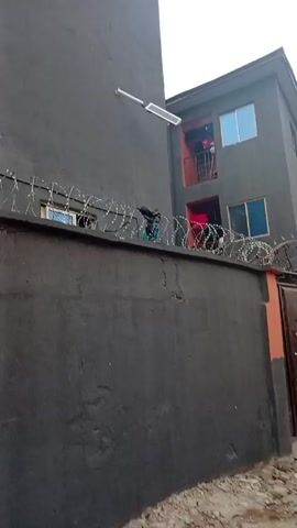 Here's a description of the video:

The video shows the exterior of a two-story building painted in dark gray. The building has windows with blue and clear glass and a balcony with red trim. On the balcony, clothes are hanging out to dry. 

A high, gray concrete wall topped with barbed wire surrounds the property. A streetlight is attached to the wall of an adjacent building. A dark metal gate with an ornate design provides access to the property. Some weeds and debris can be seen at the base of the wall.