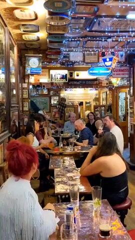 A lively scene unfolds within a dimly lit, decorated pub. The ceiling is adorned with numerous vintage signs and strings of fairy lights, creating a festive atmosphere. Several people are seated at tables, some engaged in conversation, while others are being entertained by live music. In the foreground, a woman with bright red hair faces away from the camera, wearing a white fringed top. To her left, a woman with dark hair plays an acoustic guitar and sings, accompanied by another musician. A group of men and women are seated at tables in the mid-ground, some holding drinks. A man in a red vest stands to the right, looking towards the musicians. The tables are covered with a shiny, reflective material. Several glasses, including a pint of dark beer, are visible on the tables. The overall impression is of a warm and convivial gathering in a characterful pub.