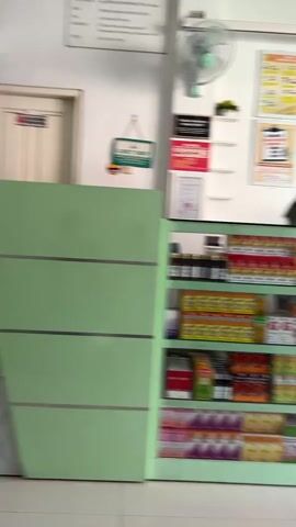 The video shows the interior of a pharmacy or clinic. A person wearing a hijab and mask is visible behind a counter filled with shelves of medicines and health products. The shelves are organized with various boxes and bottles. To the right, a glass door with a green logo and the words "KLINIK SEKAR" is partially open, leading to an outdoor area. A fan is mounted on the wall above the counter. The overall environment appears clean and functional, typical of a healthcare facility.