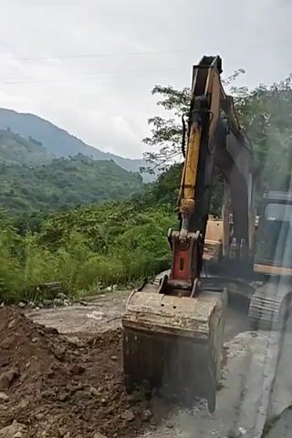 The video shows an excavator digging on a mountainside. The excavator is yellow and has a large bucket. The bucket is filled with dirt and rocks. The excavator is digging on the side of a road that is under construction. The road is narrow and appears to be in a rural area. The mountains are covered in trees and vegetation. There are some houses visible in the distance on the mountainside. The sky is overcast. The video is taken from a close-up perspective of the excavator's bucket.