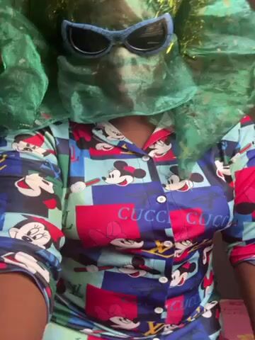 A person is shown in a video wearing a matching pajama set. The pajamas are predominantly blue, red, and green, with a cartoon pattern. The person has on dark sunglasses and a green net covering their head and face. The video is taken in a room with white and grey walls. The person stands centered in the frame and moves their arms.
