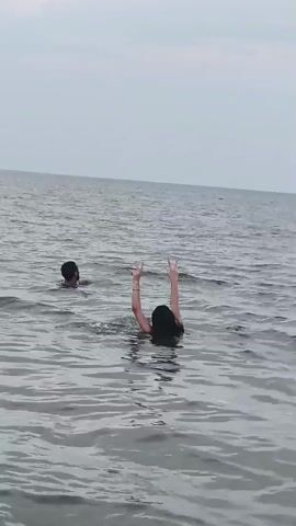 The video shows two people swimming in the ocean. In the foreground, a person with dark hair is facing away from the camera, with their arms raised and hands in a peace sign gesture. In the background, to the left, a person with dark hair and a beard is swimming towards the camera, smiling and making a wave gesture. The water is choppy and the sky is overcast.