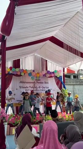 A group of young children are performing a dance routine on a stage. They are dressed in casual clothing, and some are wearing hats. The stage is decorated with balloons and banners. A tent is set up over the stage, with red and white draping. An audience of people, mostly women in headscarves, are seated in front of the stage. The event appears to be a school performance or graduation ceremony. The backdrop of the stage has text that reads "AKHIRUSSANAH, PENTAS SENI & WISUDA TAHFIDZ AL-QUR'AN SDIT AZ-ZAHRA CURUG" followed by dates and locations. The overall atmosphere is festive and celebratory.