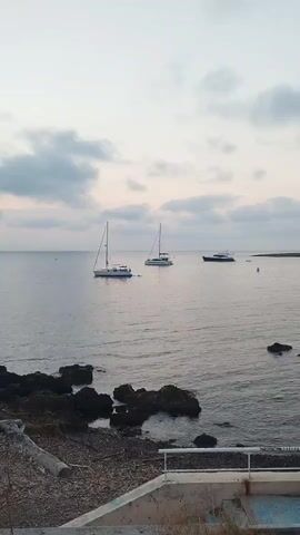 The video shows a serene coastal scene at dusk. Three boats are anchored in the calm water. Two are sailboats with their sails down, and one is a larger motor yacht. The shoreline in the foreground is rocky, with dark, jagged rocks partially submerged in the water. A concrete structure, possibly a retaining wall or part of a building, is visible at the bottom of the frame, with a set of steps leading down. The sky is overcast with soft, diffused light, indicating either sunrise or sunset. The overall atmosphere is peaceful and tranquil. There are no people visible in the video.