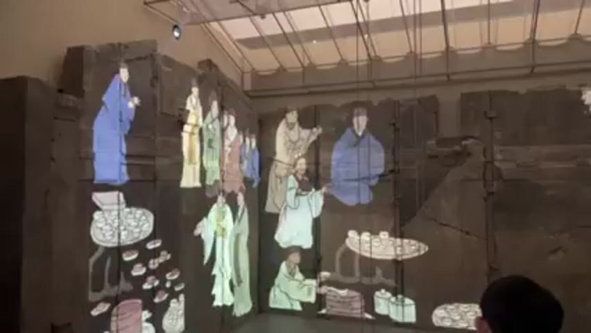 The video shows a museum exhibit where a large, dark, stone wall is being used as a canvas for a colorful animated projection. The projection depicts a traditional Chinese scene, with figures in robes, tables set with tea cups, and what appears to be food. The wall is cracked and appears to be ancient. A person is standing in the foreground, viewing the exhibit. The lighting is dim, with a few spotlights illuminating the scene. There are visible beams or wires on the ceiling above. The space appears to be indoors.