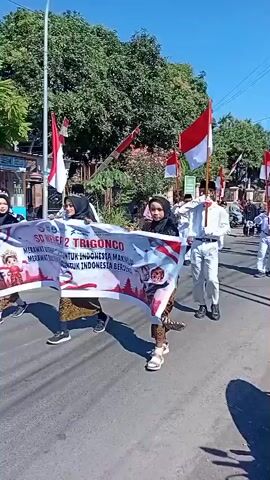 A group of people, mostly children and young adults, are participating in a parade. Many are carrying Indonesian flags, which are red and white. Some are wearing white uniforms with red accents, and others are wearing more casual clothing. One person in the foreground on the left is holding a banner with text and an image of a child on it. The parade is taking place on a paved road with trees and buildings visible in the background. The sky is clear and blue. The overall atmosphere appears festive.