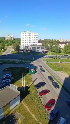 The video is an aerial view of an urban or suburban environment on a sunny day. In the foreground, there are buildings and a parking lot with several cars. Further back, there is a road with more cars and a person riding a bicycle. There are also trees and green spaces, as well as taller buildings in the distance. The scene appears to be a typical day in a residential or mixed-use area.