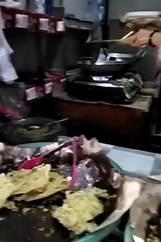 A woman wearing a grey hijab and a pink shirt with a grey apron is cooking at a stove. She is using a spatula to stir food in a wok. The stove is on, and there is a blue flame visible. In the foreground, there are plates of food wrapped in paper and banana leaves. The environment appears to be a kitchen or food stall.
