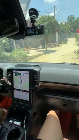 A dashboard-level point-of-view video shows a car driving down a narrow street. The car appears new, with a large GPS screen, a dashcam affixed to the windshield, wood trim, and red floor lighting. The car passes buildings and a Vietnamese flag, and there are power lines overhead. The driver is visible, wearing shorts, sandals, and a medical brace. Music plays in the background.