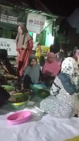 The video shows a group of people, mostly women wearing hijabs, gathered on the floor. They appear to be sharing a meal or preparing food, with several bowls and plates visible. The setting seems to be outdoors at night, with some artificial lighting illuminating the scene. In the background, there are banners and structures that suggest a community event or gathering. Some individuals are actively engaged in serving or eating, while others are observing or conversing. The atmosphere appears to be casual and communal.