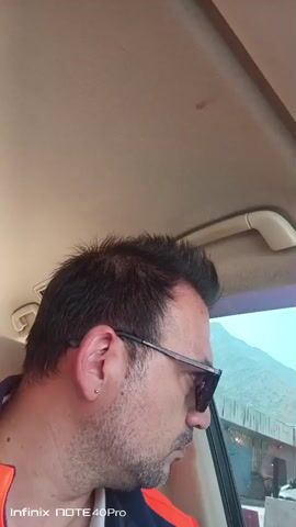 A man wearing sunglasses and a blue and orange jacket sits in the passenger seat of a car. He is looking down and to the right. The interior of the car is visible, including the headliner and a portion of the dashboard. Outside the car, a mountainous landscape and some buildings can be seen. The "Infinix NOTE 40Pro" watermark is visible in the bottom left corner.