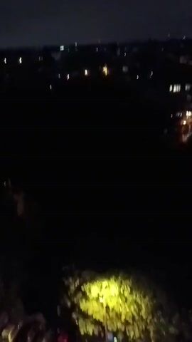 The video captures a nighttime scene from an elevated perspective, possibly from a balcony or window. It begins with a view of a parking lot illuminated by yellow lights, with several cars parked in designated spots. Trees and foliage are visible around the parking area. As the camera pans and tilts, the view shifts to a darker landscape with more trees and some distant lights that suggest a town or city. There are no people clearly visible in the footage, nor are there any specific actions or interactions depicted. The overall impression is a quiet, dimly lit environment at night.