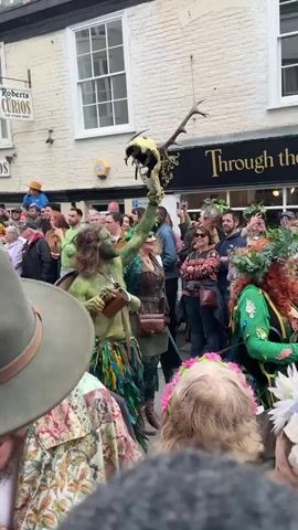 A crowd of people is standing in front of a building called "Through the looking glass", watching a man dressed in green body paint. He has long hair and is holding a skull with antlers in one hand and a wooden mug in the other. He is also carrying a shield and has a book in his hand. Some people in the crowd are taking pictures with their phones, and some are wearing flower crowns or green clothing. The event seems to be festive, with people cheering.