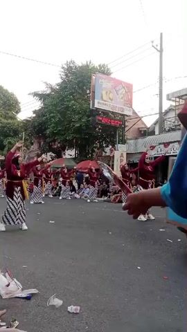 A group of people, appearing to be mostly women and girls, are performing a dance in the middle of a street. They are dressed in traditional clothing, which includes maroon tops, patterned skirts, and headscarves. Some are wearing white sneakers. They are all facing the same direction and their arms are raised in synchronized movements. In the background, there are spectators sitting on the side of the road, some trees, buildings, and a large billboard with advertisements. The ground is paved and there is some litter scattered around. The lighting suggests it is daytime.