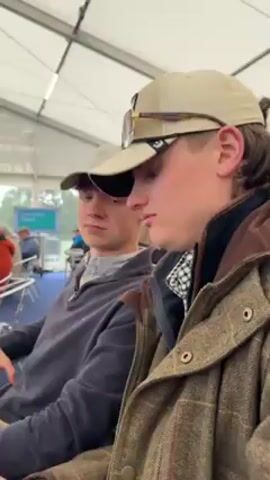 Here is a description of the video:

The video shows a scene inside of a tent, with a crowd of people. The camera moves around, focusing on various individuals and groups. Initially, the focus is on two young men seated beside each other, one in a tweed jacket and a baseball cap with sunglasses perched on top, the other in a similar cap and a gray sweater. The camera then pans to show a group of men standing and talking, including one in a pink shirt. 

Subsequently, the camera focuses on a young man wearing round sunglasses and a blue vest over a pink shirt. It then shows the scene more widely, revealing a large crowd of people seated at tables under the tent. The tables are covered in plastic cups, and many of the people are looking out towards the view outside. The video ends with an overall shot of the crowd and the view beyond the tent.
