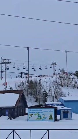 The video presents a daytime view of a ski resort covered in snow under an overcast sky. A ski lift carrying skiers is visible on the hillside. A wooden building with a snow-covered roof is situated in the foreground, next to a sign that features a winter-themed illustration with text in Russian. Snow groomers are lined up in front of the building. Skiers can be seen descending the slope. Electrical wires stretch across the top of the frame.