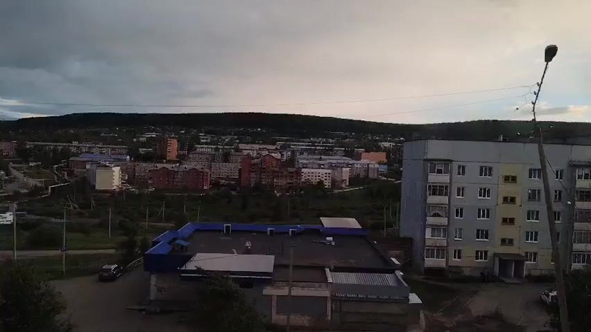 An outdoor, daytime, aerial shot showcases a somewhat overcast sky above a small town. In the foreground, a light-colored van is parked on the left side. To the right, a multi-story apartment building dominates the frame. A flat-roofed commercial building, painted blue and white, is situated in the middle ground. The background reveals more residential buildings and a tree-covered hill. The overall impression is one of a quiet, possibly remote, community.