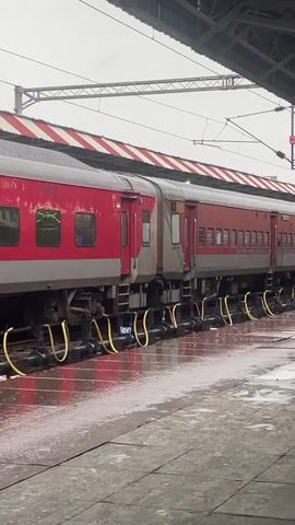 The video shows a stationary red and grey passenger train at a covered station platform. The platform is wet, indicating recent rain or cleaning, with puddles visible. Yellow hoses are attached to the underside of the train cars. The sky is overcast. A person is visible in the background walking on the platform.