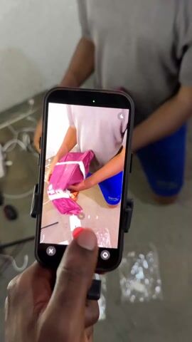 A person holds a smartphone in front of them, recording a video.  The phone screen shows another person wearing a grey t-shirt and blue shorts, opening a purple and white backpack on the floor. The camera is close, showing the hands of the person opening the backpack.  The floor is concrete and has some white cables, a black box, and plastic packaging scattered on it.  The phone is mounted in a black holder. The camera records the person pulling out contents from the backpack.