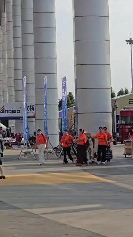 At 00:00, a group of people in orange shirts and white pants are walking around an outdoor event space. They are near large white pillars and several blue banners. One person in an orange shirt is pushing a cart. At 00:01, the group continues to move. A truck with "NEXT ERA" written on it is visible in the background. At 00:02, more people are seen walking, some in orange shirts, others in casual clothing. At 00:03, the group of people in orange shirts are gathered near the truck. A person in a black shirt and white pants walks away from the camera. At 00:04, the scene shows more of the event space with banners and people. At 00:05, the view shifts slightly, and a blue banner on a tripod is prominent in the foreground.