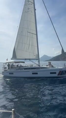 A white sailboat, identified by the text "CH2" and "Ancyra Sailing" on its main sail, is seen cruising on clear blue water under a bright, sunny sky. Several people are visible on board, some seated on the deck under a canopy, and at least one person near the bow. The boat features a red, white, and blue stripe pattern on its hull near the stern. A lush, green hilly coastline forms the backdrop, suggesting a coastal setting. The water shows ripples from the boat's movement as it glides along. The scene depicts a serene daytime sailing experience in a location consistent with Luštica, Montenegro.
