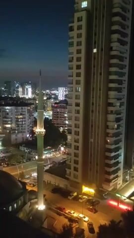 The video shows a night view of a city from a high vantage point. In the foreground, there is a minaret of a mosque, illuminated by lights. Adjacent to the minaret is a large, modern apartment building with many windows. Below, a street is visible with parked cars and streetlights. In the background, other city buildings and lights can be seen, indicating an urban environment at dusk or night. The sky is dark blue.