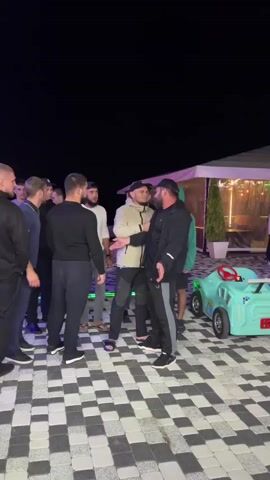 A group of men are standing on a checkered outdoor patio at night. One man in a light-colored jacket is holding a microphone and speaking to another man in a black jacket. The other men are observing the interaction. In the background, there is an illuminated tent and a child's toy car. The lighting suggests it is evening or night.