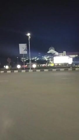The video shows a nighttime view of a building with a dome, illuminated by lights. There are several streetlights and decorative lights along a road in the foreground, which is paved and has white markings. A billboard with a black and white image is visible on the left side of the frame, mounted on a pole. The sky is dark, indicating it is nighttime. The camera appears to be mounted on a moving vehicle, as evidenced by the slight motion blur and the changing perspective of the scene.