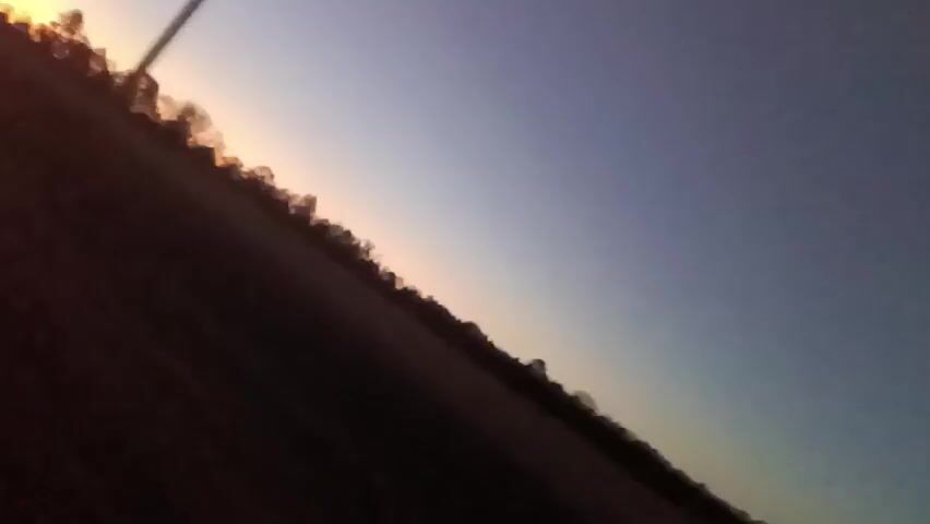 The video shows a blurred view of trees and a sky transitioning from orange to blue, possibly at sunset or sunrise. The camera angle is tilted. A dark contrail is visible diagonally across the sky. Mechanical sounds are audible.