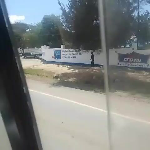 A car is traveling down a paved road past "The Milimani School," in what appears to be a warm, sunny climate. Trees line the side of the road.  The school is a white, walled compound with a gate.  People and vehicles are seen near the school's entrance.  Advertisements for paint are painted on the exterior of the compound's walls.  The car continues driving past the compound, with more paint advertisements visible.  Other vehicles pass the car.