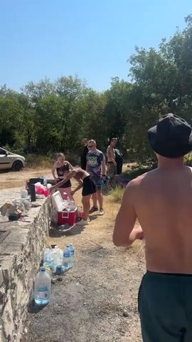 A group of people are gathered outdoors, preparing for or enjoying a picnic or barbecue. The scene appears to be in a rural or park-like setting with trees and a clear blue sky. Several individuals are visible, some standing around a stone counter with food and coolers, while others are walking or standing nearby. There are at least two cars visible in the background, one a silver hatchback and another a darker colored van. A barbecue grill is set up on a stone structure, and bottles of water are scattered on the ground. A blue umbrella is also visible, providing shade. The overall impression is a casual outdoor gathering on a sunny day.