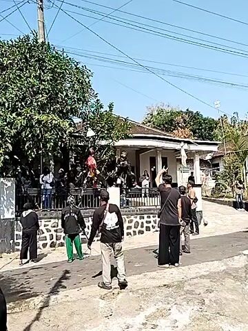 A group of people are gathered on a street in front of a house. Some people are standing on a raised platform in front of the house, while others are on the street. One person is dressed in a costume that resembles a horse. There is a tree to the left of the house. The sky is blue and clear. The scene appears to be a street performance or celebration.