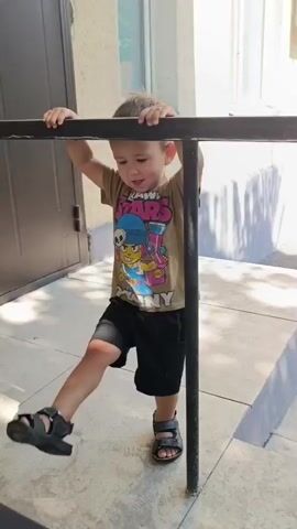 A young boy with light brown hair and wearing a tan t-shirt with a Brawl Stars Penny graphic and black shorts stands outdoors, holding onto a black metal railing. He is wearing black sandals. The boy is positioned on a tiled surface in front of a building with light-colored walls and windows. To his right, a concrete step leads upwards. The background shows greenery and a blurred figure in the distance. The boy shifts his weight and looks towards the camera.