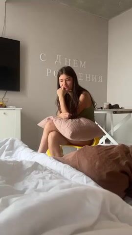 A still, medium shot captures a bedroom scene featuring a young woman sitting on a yellow chair near the foot of a bed. She's wearing a green tank top and holds a pink pillow in her lap. Her dark hair is long, and she rests her chin in her hand with a thoughtful expression, her gaze directed slightly downward. The bed is unmade, with white sheets and a brown blanket bunched at the end. On the wall behind her, white lettering spells out "С Днем Рождения" (Happy Birthday in Russian). A black television hangs on the wall to the left, above a white cabinet. To the right of the woman is a white desk with various objects on it. The room is brightly lit, and the overall impression is one of quiet introspection.